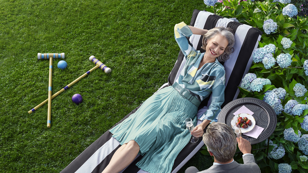 Gray haired woman in blue relaxes on black/white chaise. Croquet mallets on grass, blue hydrangeas behind.