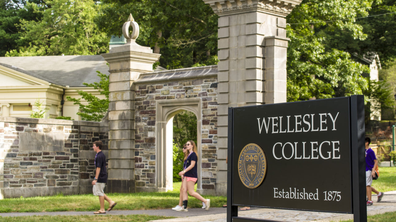 Young women walk through gate by Wellesley College sign.