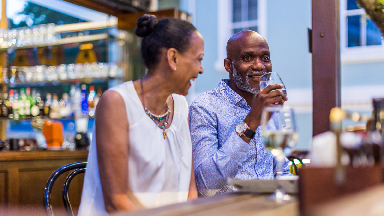 Black woman in white, man in blue shirt drinking white wine at bar